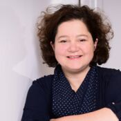 Eine Frau mit braunen Haaren und einem gepunkteten Halstuch lächelt in die Kamera.