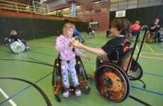 Ein Mädchen im Rollstuhl gibt einem Jungen im Rollstuhl einen gelben Tennisball