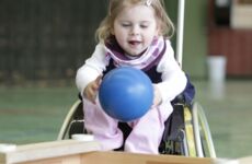 Ein kleines Mädchen im Rollstuhl greift nach einem blauen Ball