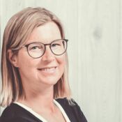 Eine Frau mit schulterlangen, blonden Haaren und runder Brille lacht seitlich in die Kamera