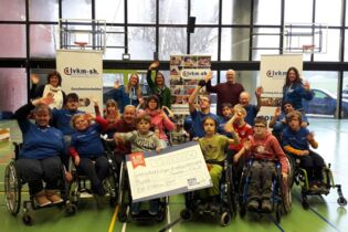 Eine Gruppe Rollstuhlfahrende freut sich über einen Spendenscheck der R.SH hilft helfen-Stiftung
